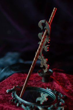 Tahtadan yapılmış, figür oymalarla süslenmiş sihirli bir değnek, eski bir aynanın yanında duruyor ve koyu kırmızı kadife arka planda işlenmiş demir bir ayakta duruyor. Sihirli dekor dikey olarak yakın çekim
