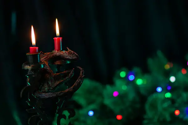 Burning red christmas candles in a handmade candlestick with a dragon figurine symbol of 2024 on the background of a sprig of a Christmas tree decorated with a garland of flashing lights, clothe-up