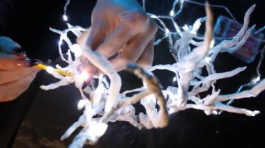 Beyefendinin elleri, tacı ince beyaz dallarla yontuyor, onlara parlak bir çelenk dokuyor mavi neon ışıkta koyu bir arka planda. Mistik bir aksesuar yapma süreci, 4k yakın plan.