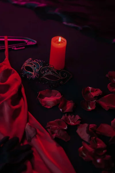 Massage candle on a dark bed among rose petals, womens trinkets, next to a red satin dress. Preparing for a passionate date on Valentines Day, close-up