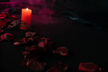 Gül yaprakları bir mumla birlikte siyah kumaşın üzerine saçılmış ve açık bir maske ile kırmızı neon ışığın dumanlı arka planına karşıdır. Sevgililer Günü kartı. Yakın plan.