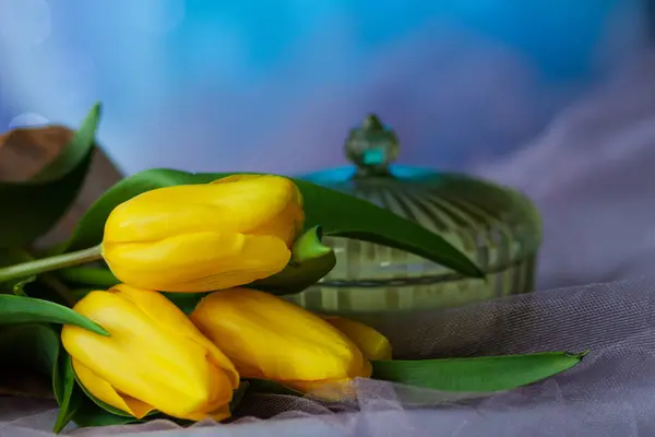 Green crystal ribbed vase with lid among yellow tulips on blue background with shining sequins close-up. Holiday Card with Spring Flowers, Greeting Text Background