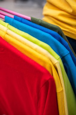 Askıda asılı bir grup sade renkli tişört. Mağazada çeşitli parlak kıyafetler, yakın çekim, dikey çerçeve
