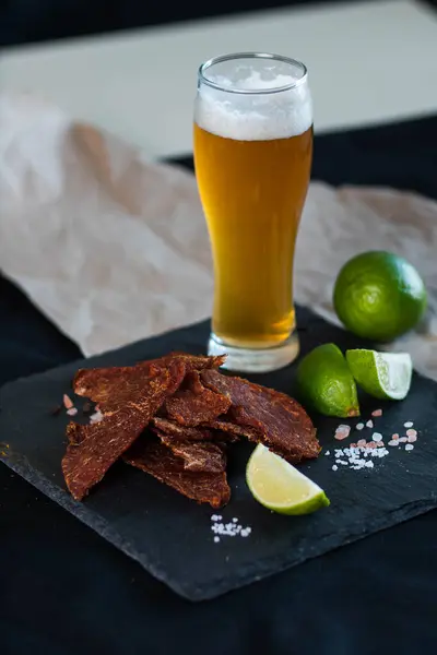 A glass of frothy light beer next to narrowed meat, jerky snacks for beer, spices, chopped lime laid out on a stone black tray. Food, Drink Close-up Vertical