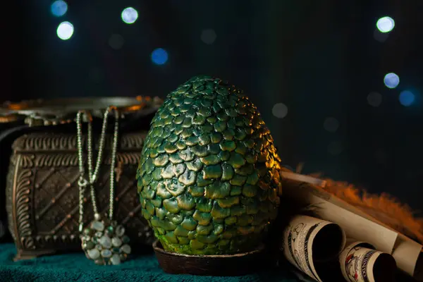 A large green scaly dragon egg stands on a stand next to an antique chest with a necklace among scrolls and fabrics, on a dark background with glowing bokeh. Fabulous treasures, close-up
