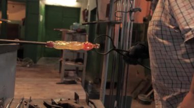 Koruyucu eldivenlerdeki ustanın elleri metal maşaları tutar ve onları demir bir boruyla savrulan erimiş cam bir figür haline getirir. Atölyede cam süsleme işlemi, yakın plan.