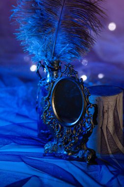 İşlenmiş demir çerçeveli eski bir ayna, parlak mavi arka plandaki bir çelengin ışıldayan ışıkları arasında devekuşu tüyü şişesinin yanında duruyor. Büyülü atmosfer, dikey yakın çekim