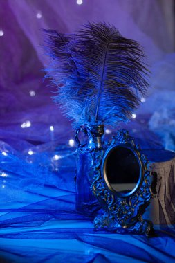 İşlenmiş demir çerçeveli eski bir ayna, parlak mavi arka plandaki bir çelengin ışıldayan ışıkları arasında devekuşu tüyü şişesinin yanında duruyor. Büyülü atmosfer, dikey yakın çekim