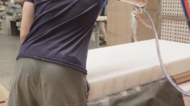 Atölyede çalışan bir işçi elinde sprey şişesiyle bir hortum tutar ve gelecekteki kanepe için yatağın üzerine su döker. Mobilya üretme süreci, yavaş çekim.