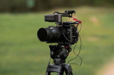 Profesyonel kamera, siyah, yeşil bir alanın arka planına karşı tripoda monte edilmiş, çekime hazır. Video ve ses kayıt ekipmanları, yakın çekim.