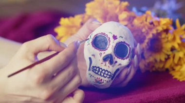 Bir el bir fırça tutar ve kafatası heykelciğini mavi neon ışıkta sarı çiçeklerin arka planına karşı renkli bir süsleme ile boyar. Meksika 'daki Santa Muerte tatiline hazırlanıyorum, yakın plan.