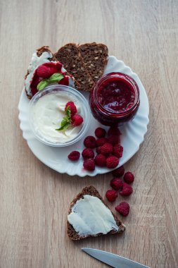 Summer breakfast on a white plate on the table with homemade white goat butter, fresh raspberry jam and slices of bread, garnished with berries. Top view, vertical, close-up clipart