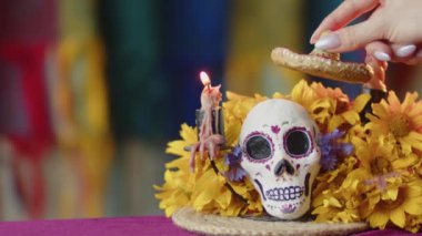 El, sarı çiçeklerin arasında boyanmış kafatası üzerine küçük zambrero ve kareli arka planda mumlar giyer. Santa Muerto Meksika aletleri, ölüm gününün sembolleri.