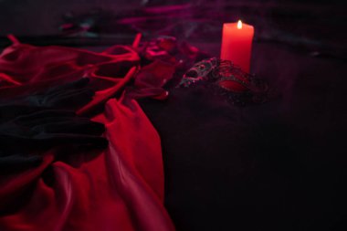Gül yaprakları, kadın ıvır zıvırları ve kırmızı saten bir elbisenin yanında, koyu renkli bir yatakta mum. Sevgililer Günü 'nde tutkulu bir randevuya hazırlanıyorum. Yakın plan.