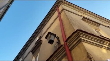 Ortaçağ mimarisinden eski bir evin boynuzu, üzerinde kırmızı bir oluğu ve sarı bir duvarda eski işlenmiş demir bir feneri olan bir sineği uçuruyor. Aşağıdaki binanın görüntüsü