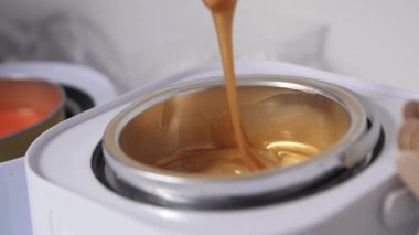 Tüy dökücü ustasının eli, sıcak metal bir kapta eritilmiş altın balmumu karışımını ahşap bir spatulayla karıştırır. Güzellik prosedürü için hazırlık, kozmetoloji yakın çekim yavaşlama