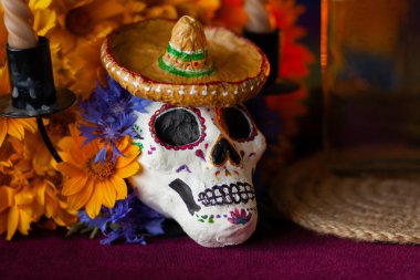 Küçük bir Lambrero 'da kafatası şeklinde boyanmış bir heykel, yanan mumların yanındaki çiçeklerle süslenir. Meksika Ölüler Günü 'nün sembolleri. Renkli, parlak bir arka planda.
