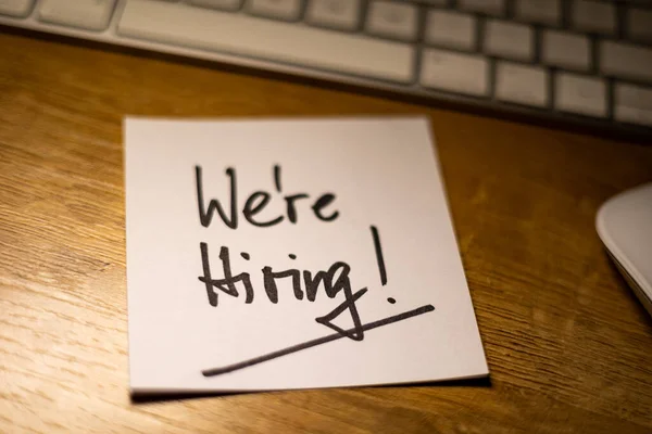 We\'re Hiring, text note. Office desk with notepad and keyboard in the background. In black the handwritten text, we are hiring. Job search, opportunity, applying and occupation.