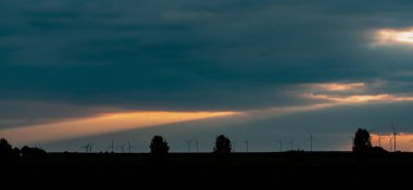 Gün batımında ufukta rüzgar türbinleri ve ağaçların silueti beliriyor. Büyük turuncu bir güneş ışığı ile karanlık bulutların arasında parlayan dramatik bir gökyüzü. Bitterfeld-Wolfen, Sachsen-Anhalt, Almanya gün batımında.