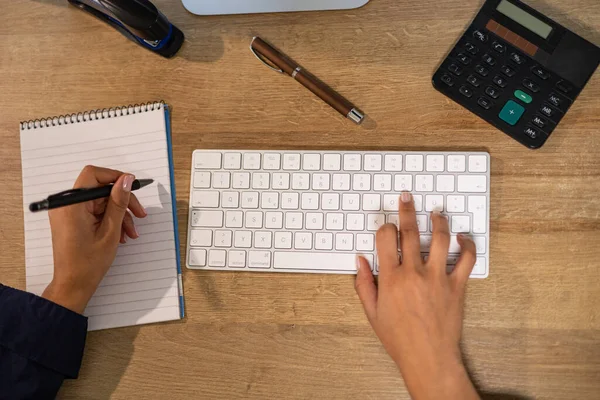 Woman types on the notebook and writes notes on a page. The office assistant hand types on the keypad and the other hand writes notes. Financial business, services or back office in the industry.