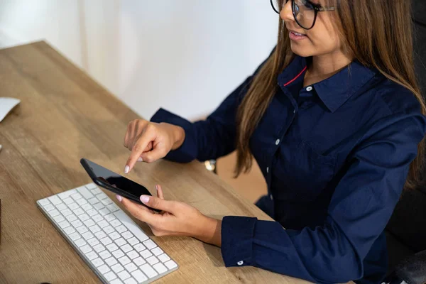 Businesswoman with glasses types on the cell phone. Woman sits at the desk, holding the cell phone and types on the display. Back office, business, file clerk, office assistance, work and management.