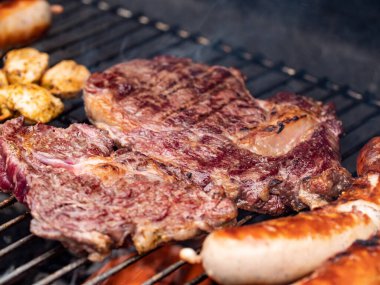 Barbeküde ızgara biftek ve sosis. Izgara bifteğe yakın çekim. Et dışarıda kavrulmuş durumda. Kömür ve ateş bifteğin altında parlıyor. Yazın barbekü..