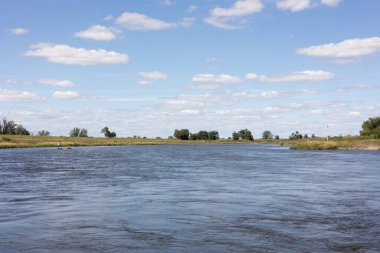Nehrin solunda ayakta duran bir kürekçi. Mavi gökyüzü, kıyı yeşil çalılar ve ağaçlarla.
