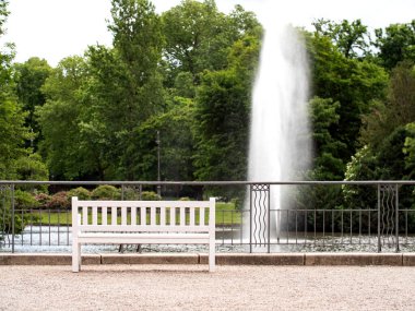 Boş beyaz park bankı. Parktaki çeşme ve ön plandaki beyaz boş park bankı. Yalnızlık ve rahatlama.