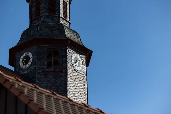 Church clock surrounded by roof shingles. Close up church tower with clock. Roff shingle tower wit ancient clock and old white, black and golden clock-face. Sunny day with blue sky and few clouds.