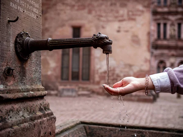 女性は古い歴史的な井戸の錆びたパイプから水の流れの下で彼女の手を保持します 城の古い歴史的井戸 — ストック写真