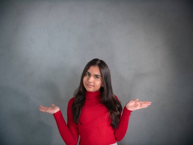 Kadın sorguluyor. Kırmızı kazaklı genç bir kadın, dirsekler bükülmüş, kollar havada ve avuçlar havada. Yüz ifadesi ciddidir, belirsizdir ve biraz tuhaftır. Gri arkaplanda izole.