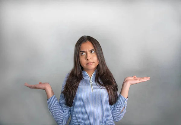 Doubting teenager, disappointed, upset Doubting young woman in blue striped jersey, elbows bent, arms raised and palms up. Questioning facial expression, the gaze is slightly downward to the left.. Isolated from the gray background.