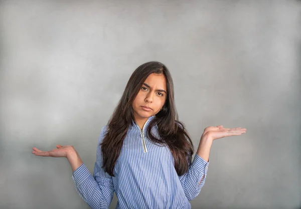Doubting young woman, disappointed, upset. Teenager in blue striped jersey, the elbows bent, arms raised and palms up. Questioning, uncertain, confused expression. The gaze is slightly downward. Isolated from the gray background.