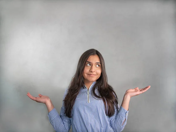 Woman thinks - Never mind! Smiling young woman in blue striped jersey, the elbows bent, arms raised and palms up. The face expression questingly and her gaze is upward to the gray background.