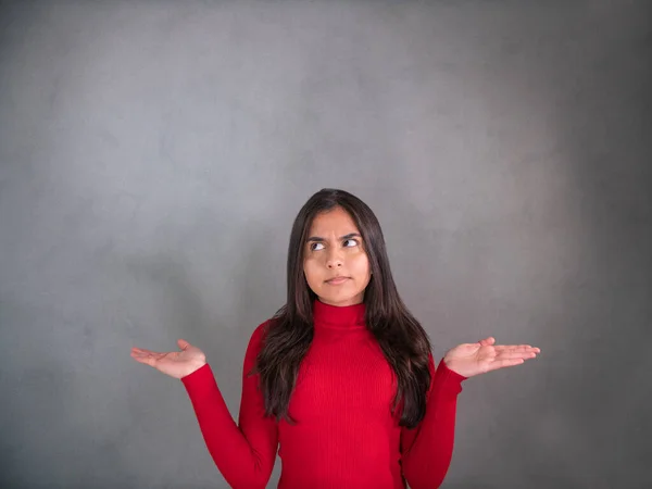 Woman thinks - What the hell! Never mind! Young woman in red pullover, the elbows bent, arms raised and palms up. The face expression is serious, uncertain and slightly questingly. She gazes to the gray background.
