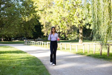 Kadın parkta yürüyor ve cep telefonuyla bir telefon görüşmesi yapıyor. Kahverengi saçlı, uzun kollu, kahve fincanı tutan bir kadın. Açık hava, halka açık park, rahat konum ve yaz. 