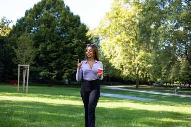 Parkta yürüyen ve akıllı telefonundan bir mesaj okuyan bir kadın. Kahverengi saçlı portre kadın, bir fincan kahve ve cep telefonu. Açık hava, halka açık park, rahat konum ve yaz. 
