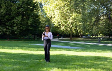Parkta yürüyen ve akıllı telefonundan bir mesaj okuyan bir kadın. Kahverengi saçlı portre kadın, bir fincan kahve ve cep telefonu. Açık hava, halka açık park, rahat konum ve yaz. 