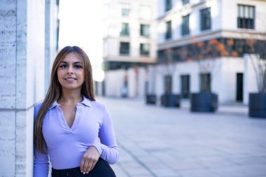 Duvara dayanmış gülümseyen kadın portresi. Kahverengi saçlı kadın şehirde halka açık bir yer. Açık hava, rahat konum ve yaz.