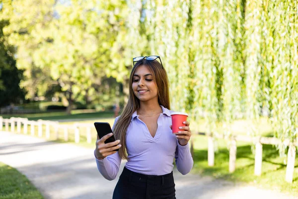 Woman walks in the park and looks at the display of her cell phone. Woman with brown hair and long sleeve holds a coffee mug while she reads a message. Outdoor, public park, relaxed position and summer.