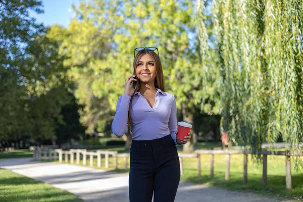 Woman walks in the park and has a phone call with her cell phone.  Woman with brown hair, long sleeve holds a coffee mug while she has a call. Outdoor, public park, relaxed position and summer.