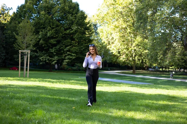 Woman walks in the park and has a phone call with her cell phone. Portrait woman with brown hair, cup of coffee and mobile phone. Outdoor, public park, relaxed position and summer.