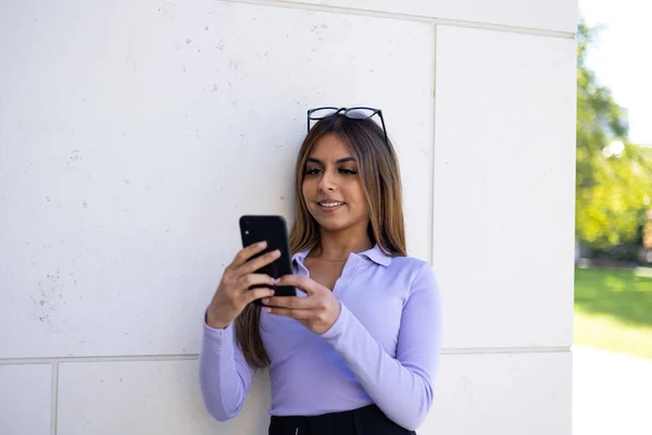 Woman in purple long sleeve shirt looks at the display of her cell phone during summer. Brown hair woman leans at the wall of the building and reads a message. In the shadow, relaxed position, glasses on top of the head.