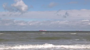 Scheveningen 'deki kıyı şeridi, Lahey' in bir bölgesi. Gökyüzünde bulutlar olan dalgalı Kuzey Denizi. Arka planda gemiler ve açık deniz rüzgar çiftliği.