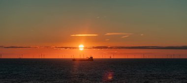 Gün batımında Kuzey Denizi 'nde bir nakliye gemisi ve rüzgar çiftliğinin silueti. Güneşin yakınında bir bulut şeridi. Scheveningen Sahili, Hollanda.