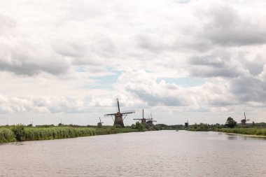 Hollanda, Kinderdijk 'te bulutlu bir günde tarihi rüzgar değirmenleri grubu. Nehir ortada, solda ve sağda sazlıklar.