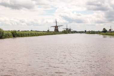 Hollanda, Kinderdijk 'te bulutlu bir günde tarihi rüzgar değirmenleri grubu. Arka planda bir tekne. Reed otları kıyıya vurur.