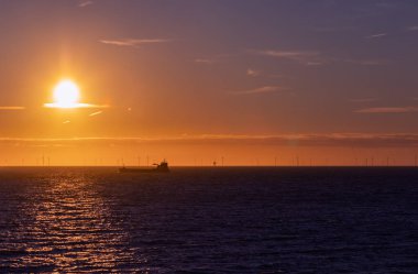 Gün batımında Kuzey Denizi 'nde bir nakliye gemisi ve rüzgar çiftliğinin silueti. Güneşin yakınında bir bulut şeridi. Scheveningen Sahili, Hollanda.