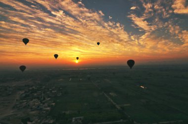 Sabahın erken saatlerinde Luxor 'da gökyüzünü kızıla boyarken bulutlarla kaplı gökyüzü, Luxor, Mısır yakınlarındaki turne şirketleri tarafından işletilen sıcak hava balonlarının gölgeleriyle kaplıyor.