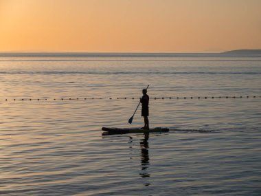 Gün batımında sakin suda yüzeye yansıyan altın rengi bir gökyüzü ile kürek çeken bir insan silueti..